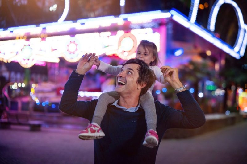Authentic shot of a happy smiling father carrying his little daughter on a shoulders having fun together in amusement park with luna park lights at night.